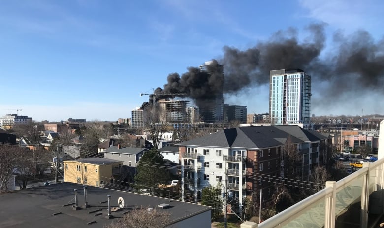 A number of city buildings are seen, with an under-construction building in the central background with large plumes of black smoke coming from it.