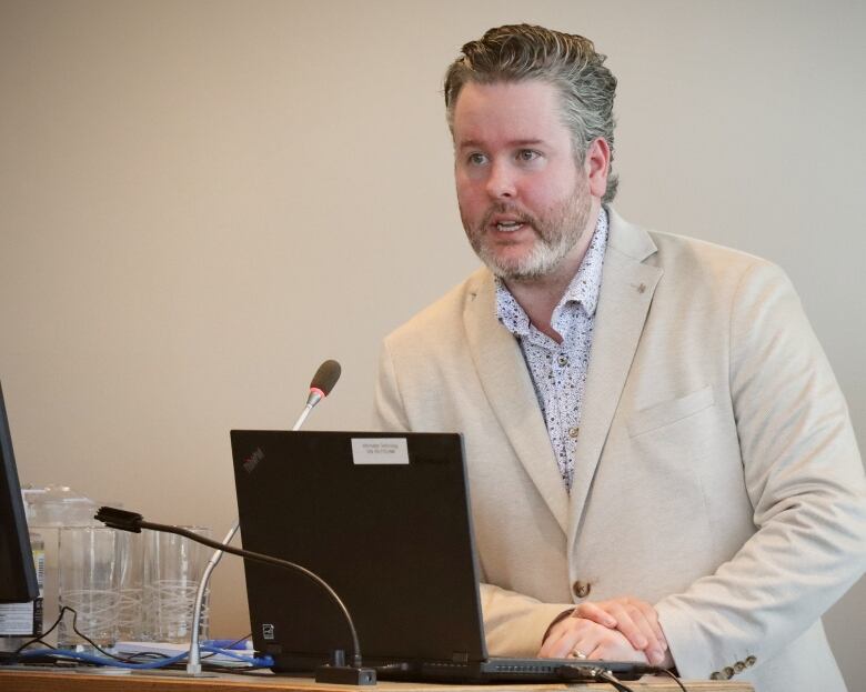 A man with a beard and moustache wearing a tan blazer with a light coloured shirt leans on a desk in front of a microphone and computer screen.