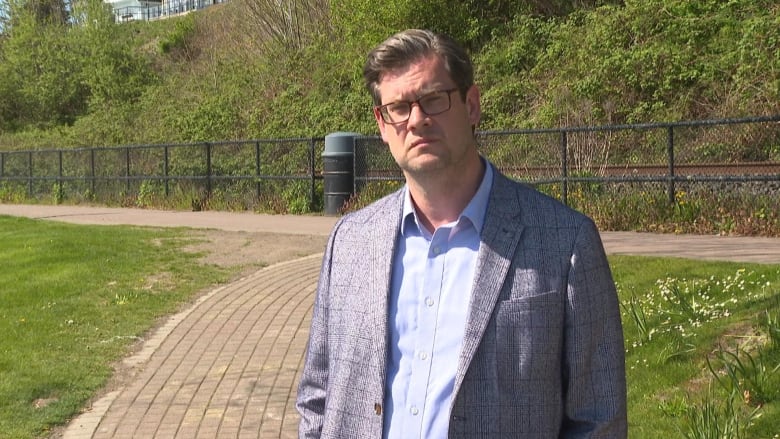 A white man wearing a grey suit-jacket stands on a grassy path.