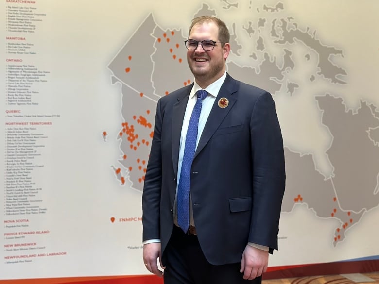 A man with brown hair and glasses wearing a blue suit white shirt and blue tie stands in front of a large grey and white map of Canada 