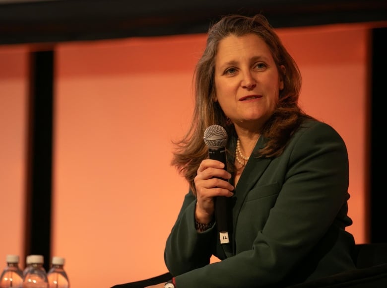 A woman with long brown hair wearing a green dress and pearl necklace sits on a stage holding a microphone. She's looking to her left and speaking. 