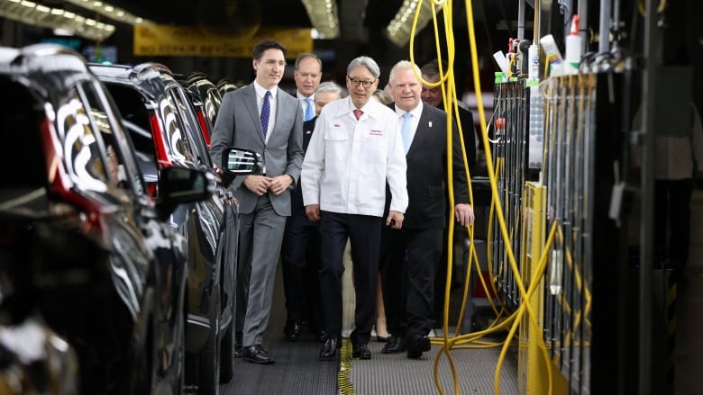 Three men walking inside a car assembly plant, with vehicles on one side and equipment on the other.