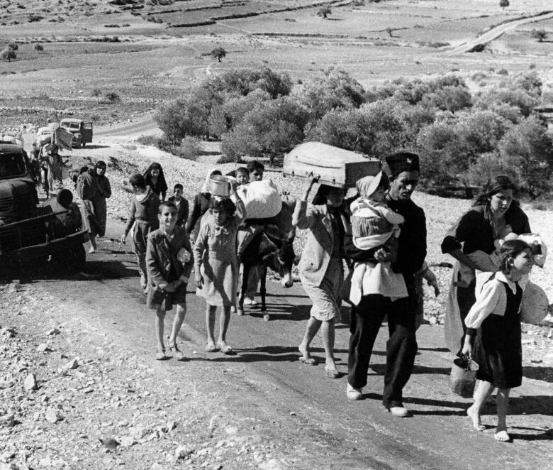 black and white photo of people walking on a dirt road carrying bags and bundles 