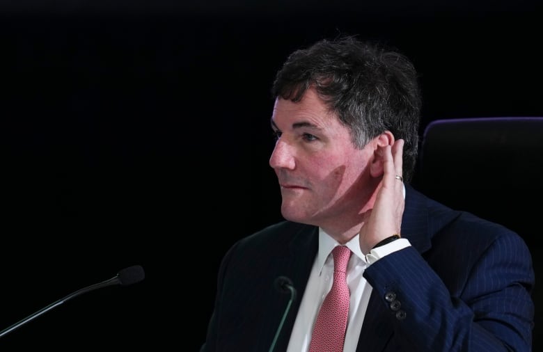 A white man wearing a red tie holds his hand to his ear while sitting in front of mics.