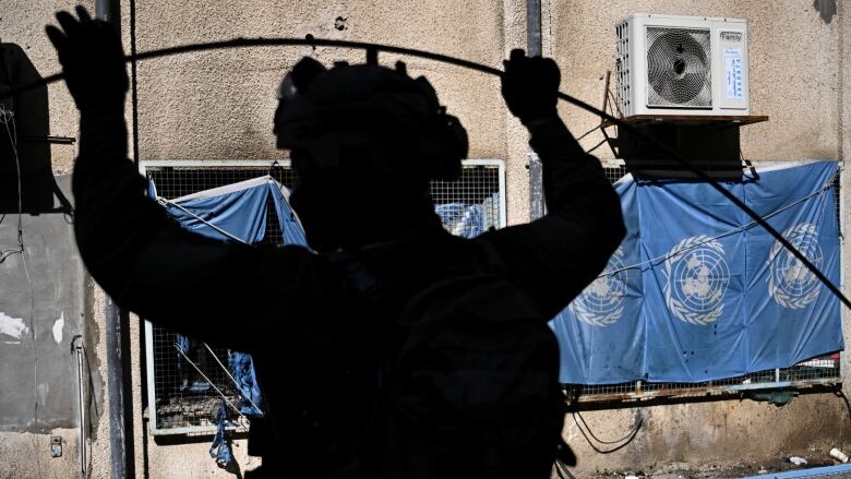 A soldier's silhouette is visible against the side of a concrete building. Blue flags for the United Nations are visible on the building.