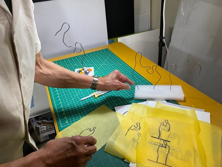 A hand points at a wire sculpture, surrounded by sheets of drawings showing the simple outline of one smaller person bending over the hand of a larger person.