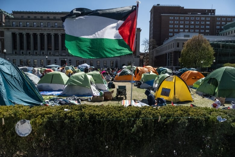 A black, white, green and red Palestinian flag waves on a pole in front of a crowd of tents on a university campus.