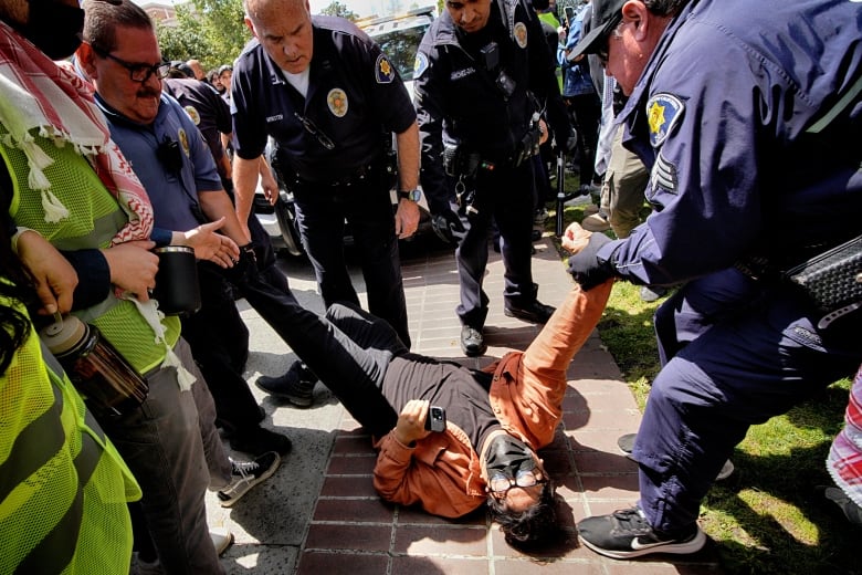 A group of security officers surround a man on the ground, with one officer on the left putting his foot on the man's body as another holds his arm. 