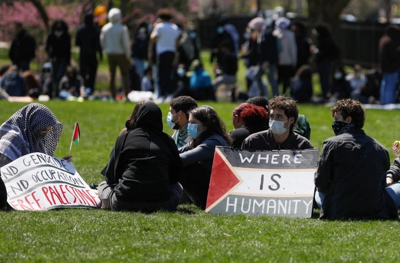 A small crowd of protesters sit on the grass with cardboard signs while a larger crowd of people stand in the background. 