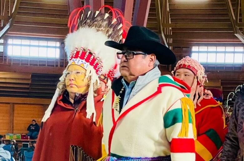 People wearing traditional regalia. On the left, a woman wears an Indigenous headress and yellow facepaint.