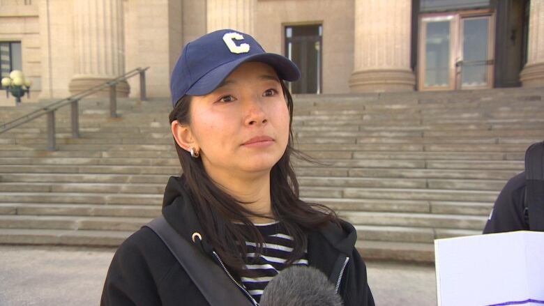 A woman in a black sweater, black and white striped shirt and blue cap is standing in front of a flight of stairs.