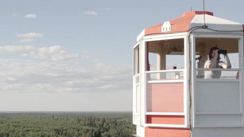 A woman in a fire tower overlooking a vast green forest. She is looking at something out of frame with binoculars.