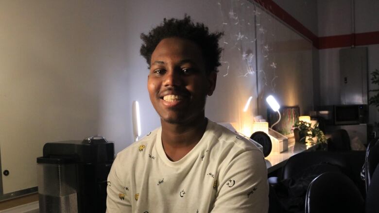 A smiling young man wearing a white, patterned t-shirt sits in a dimly lit room.