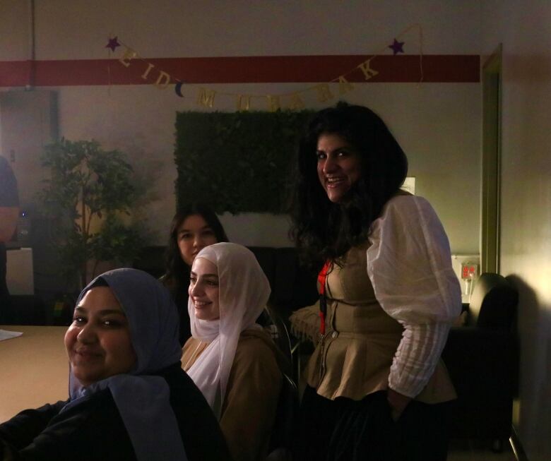 A woman is pictured standing behind her students, who are seated at a table in a dimly lit room.