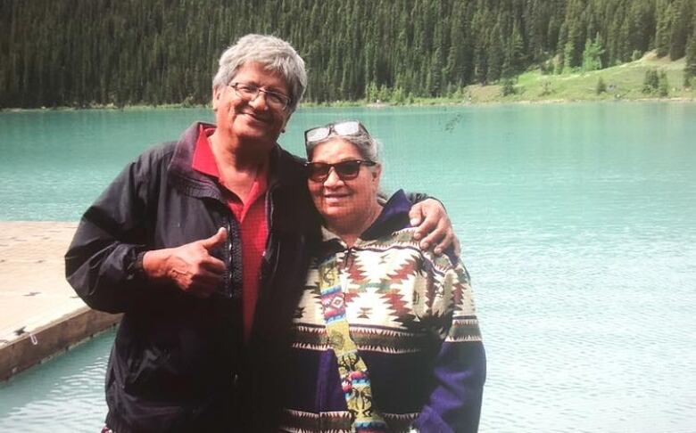 A woman wearing a long sweater and a man wearing a dark jacket pose in front of a lake of water.