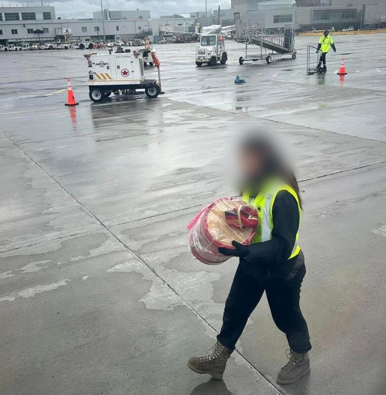 A person in a yellow vest carries a barrel-shaped decorated container wrapped in plastic. Baggage handling equipment can be seen in the background.