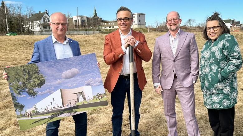 Three men in suits and a woman in a winter parka stand in a grassy field holding a shovel and an artist's rendering of what a new building could look like.