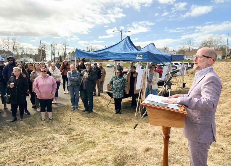 A man with a beard and moustache wearing a mauve suit speaks at a wooden podium outside on the edge of a grassy field next to a concrete parking lot.