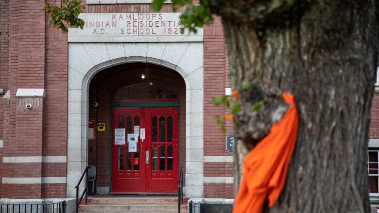 An orange ribbon is tied outside the entrance to a building which reads 'Kamloops Indian Residential A.D. School 1923'.