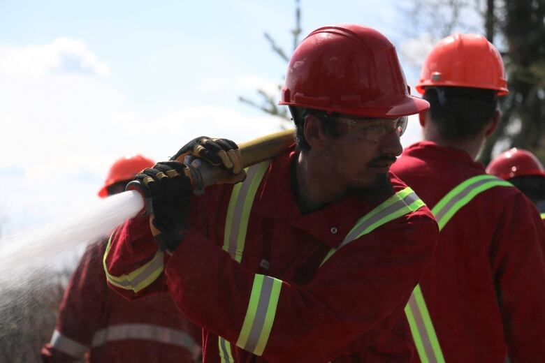 A man with a goatee holds a firehose over one shoulder as it sprays water into the bush. He's wearing a red jumpsuit  and hard hat.