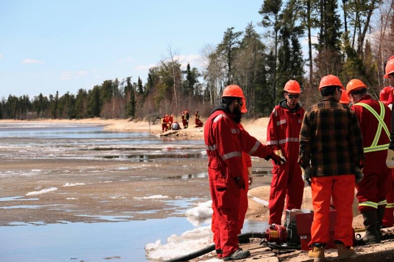 Two groups of men stand on the shores of a half-frozen lake. There are chunks of ice floating on the surface. They're putting small motors into the water to grab water from the lake.