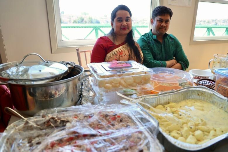 A man and a woman sit behind a table filled with Bangladeshi treats.