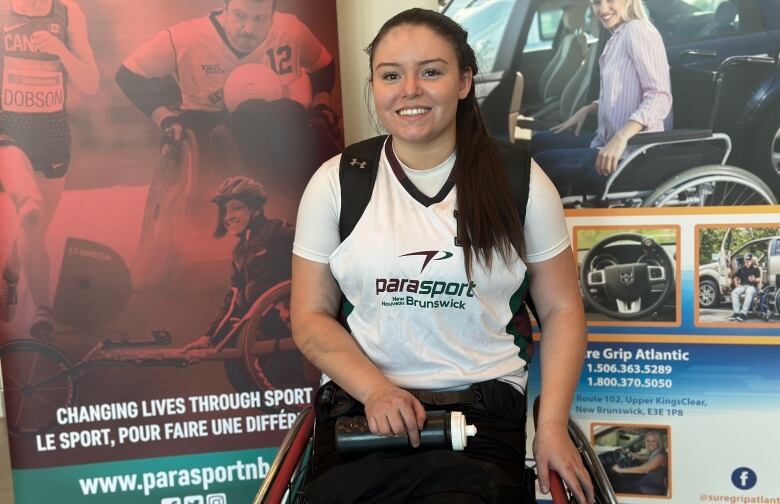 A woman with long dark hair wearing a white sports shirt sits in a wheelchair smiling.