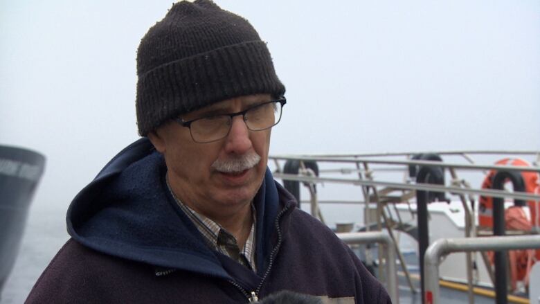 An older man wearing a black jacket and black winter hat stands on a wharf near a harbour. 