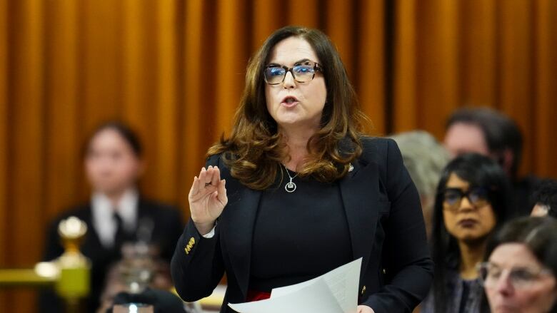 A woman in a black suit motions with her hand as she speaks in the House of Commons.