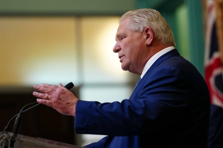 A man in a blue suit stands at a podium and motions with his hand as he speaks into a microphone.