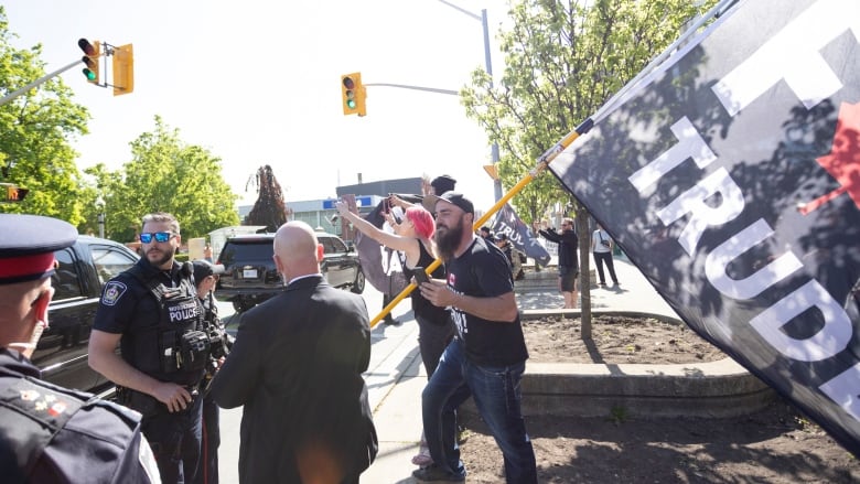 Police stand in front of a bearded man waving a large flag - the text, 'F--k Trudeau,' is only half visible within the image. 