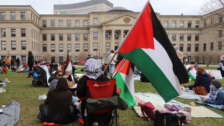 Pro-Palestinian supporters sit or stand on a lawn facing a school building at the University of Ottawa. One person with his back to the camera is wearing a keffiyeh and holding a Palestinian flag.