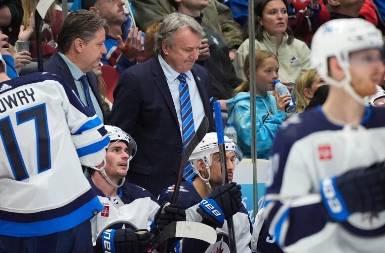 A man in a suit surrounded by Winnipeg Jets players in the foreground and fans in the stands in the background.