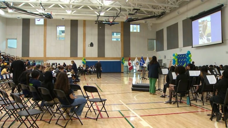 A school gymnasium is filled with people sitting on fold out chairs. A projection screen hangs down at the front of the room. 