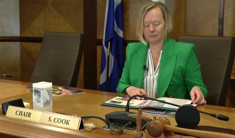 A woman in a green blazer flips through a leaflet at a wooden desk. 