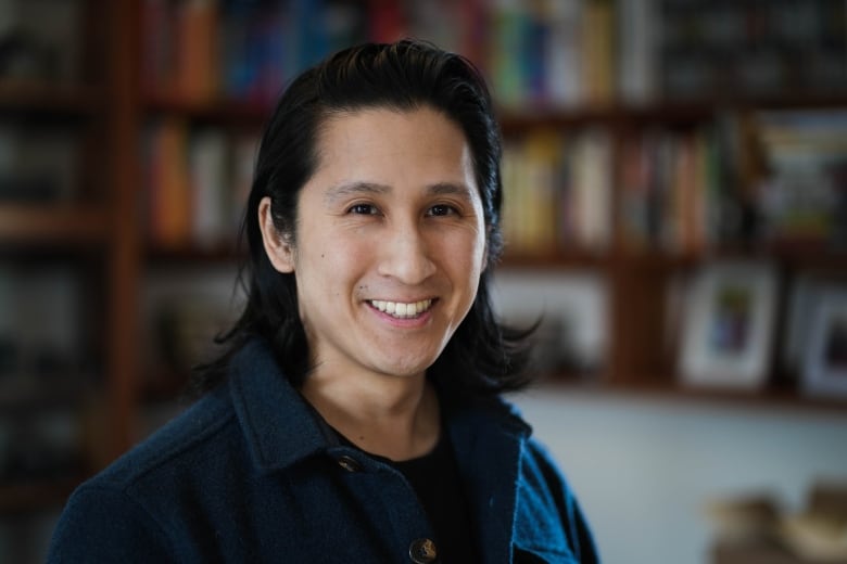 An Asian man with shoulder length dairk hair and a blue shirt stands smiling at the camera. Behind him are wooden shelves full of books and photos. 