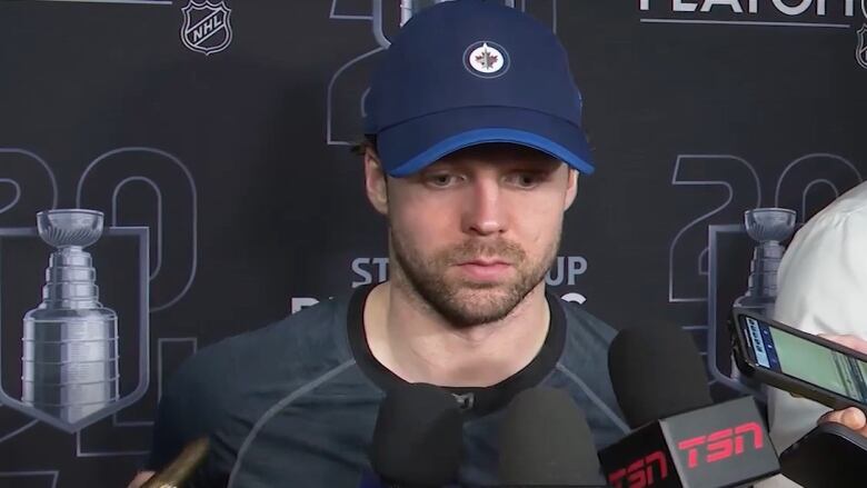 A man in a baseball cap looks down as he speaks into a microphone held by a reporter.