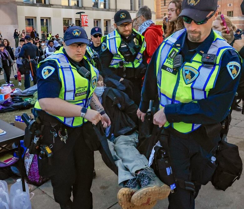 Three police officers wearing baseball caps carry a person wearing boots in an outdoor photo.