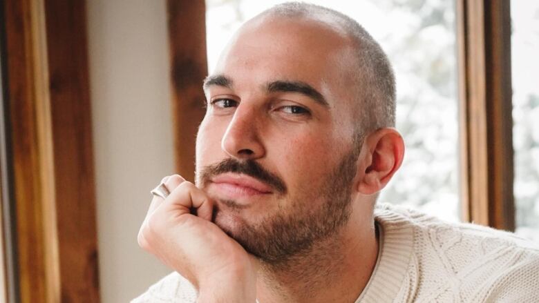 A man with short hair and a beard wearing a cable knit beige sweater with his chin resting on his hand.