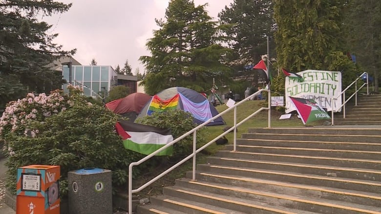 Palestinian flags are tied to the railings of the stairs, and behind them, a tent bearing a rainbow and trans flags is visible. To the right is a banner that says 'Palestine Solidarity Encampment.'