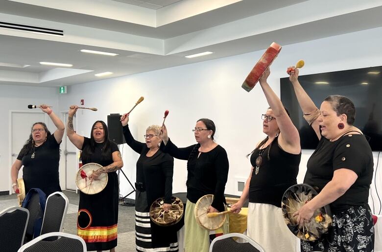 Six women wearing black tops stand together holding drums, their arms raised in song.