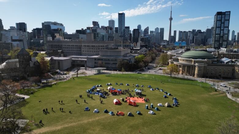 Tents arranged in a circle are seen on a plot of green space in the city, with people outside. 