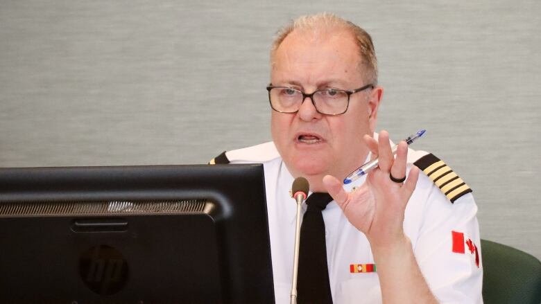 A man with thinning hair and glasses wearing a white uniform shirt with black and gold epaulettes and a dark tie speaks into a microphone and gestures with a pen in hand.