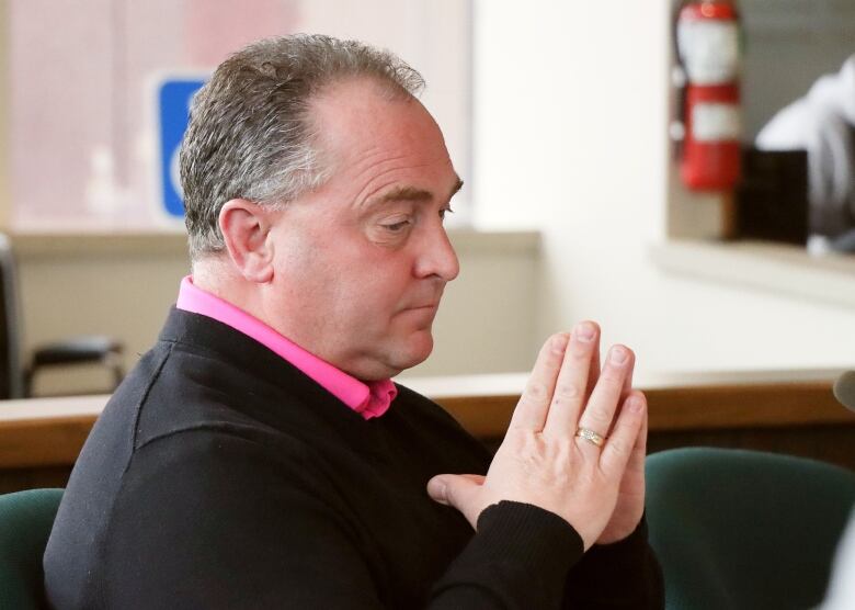 A man with greying hair, black sweater and pink shirt makes a steeple out of his hands.