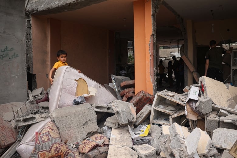 A young boy stands behind a mattress atop a pile of concrete rubble and debris inside a destroyed home. 