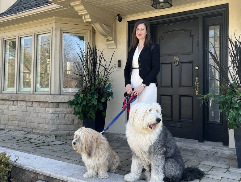 A lady with a black sport coat and white dress shirt stands outside of her beige coloured home with one large fluffy dog and one small fluffy dog