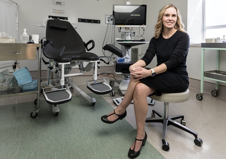 Dr. Erin Brennand sits in a clinic room. An examination bed and computer monitor can be seen behind her