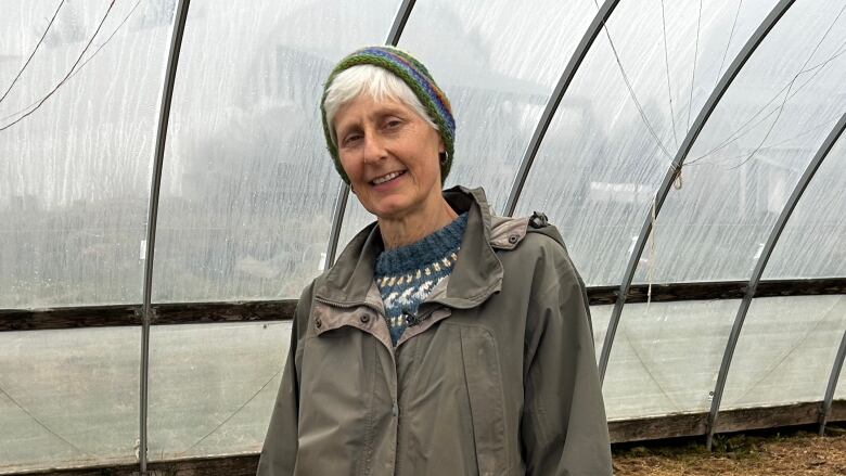 A woman wearing a large, green raincoat and rubber boots stands in a greenhouse, a large black dog at her side.