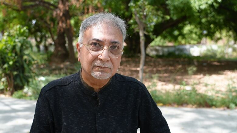 A man with grey hair, black shirt and glasses looks at the camera with trees in the background. 