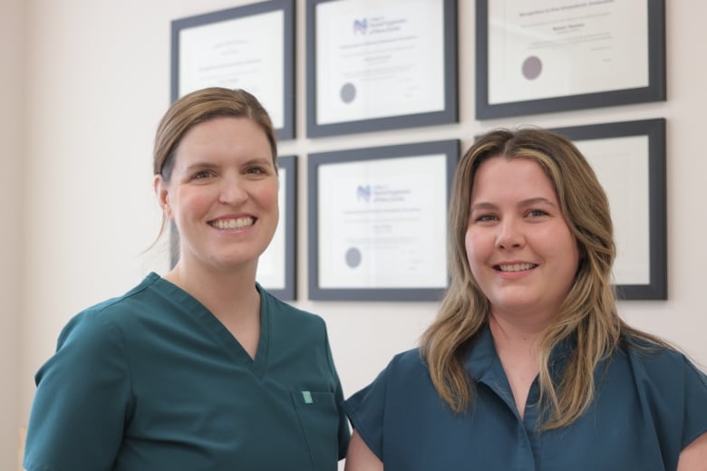 Two dental hygienists in scrubs are smiling and standing in front of framed diplomas.